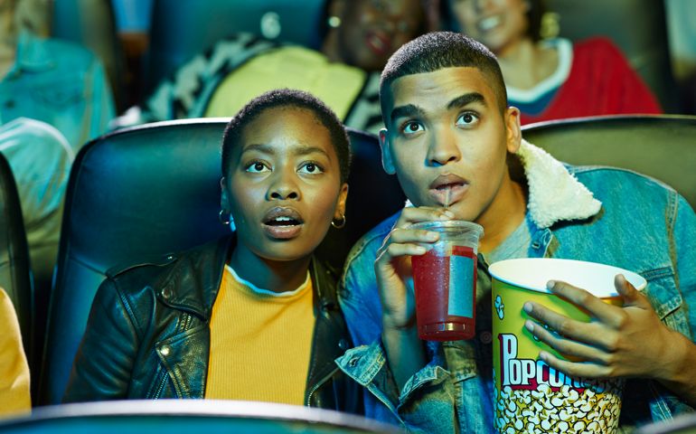 Surprised young man drinking soda while watching movie with friend in cinema hall