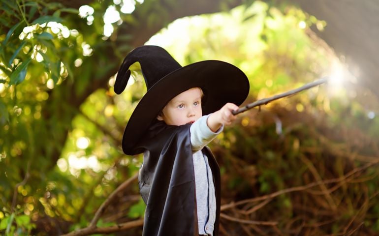 Little boy in pointed hat and black cloak playing with magic wand outdoors. Little wizard.