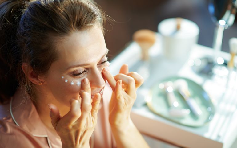 elegant female in pajamas at modern home in sunny winter day applying eye cream.
