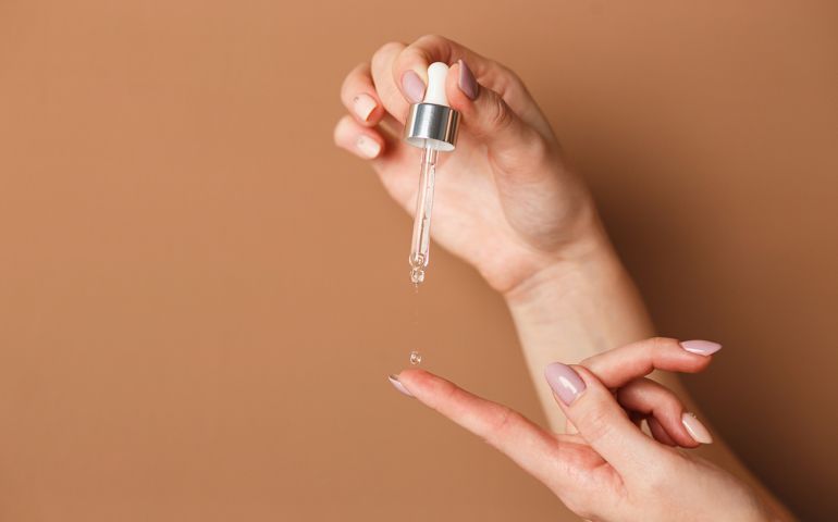 Hands of cropped white woman holding cosmetic serum pipette on the orange background.