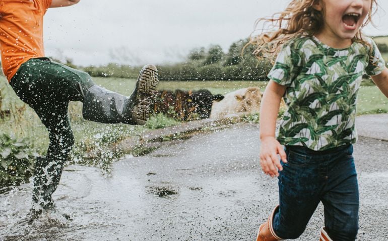 Two cute kids jump in a huge puddle, wearing welly boots. Boy playfully kicks water towards girl as she happily screams.