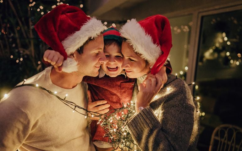 Photo of a family with one child wrapped in Christmas lights, celebrating holidays on the balcony of their apartment;