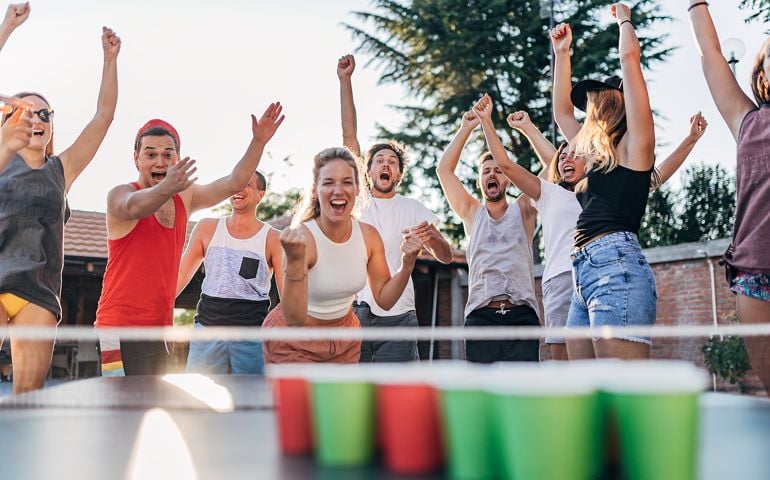 A group of young people playing beer pong in a backyard on a table tennis table on a sunny day. Young people having fun