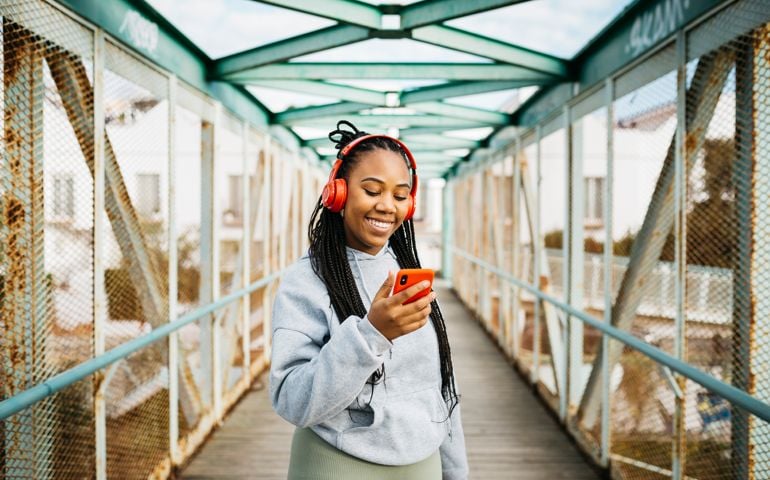 Portrait of a young smiling woman, with braided hair and sportswear, using a red smartphone and headphones, after workout on a urban bridge