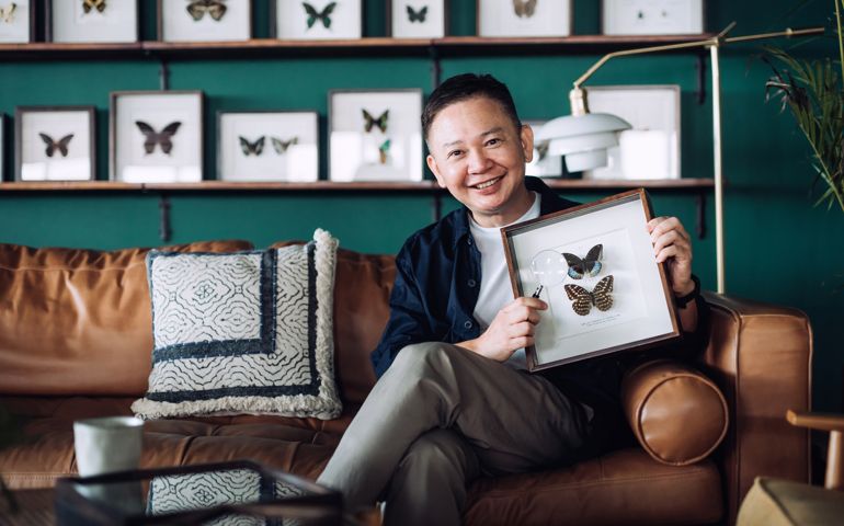 Portrait of cheerful senior man relaxing on sofa at cozy home, holding insect collection specimen holder with