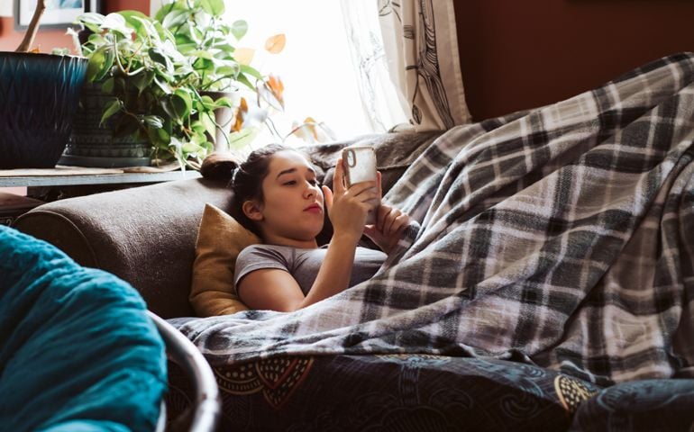 Teenage child lying on couch under a blanket watching something on phone.