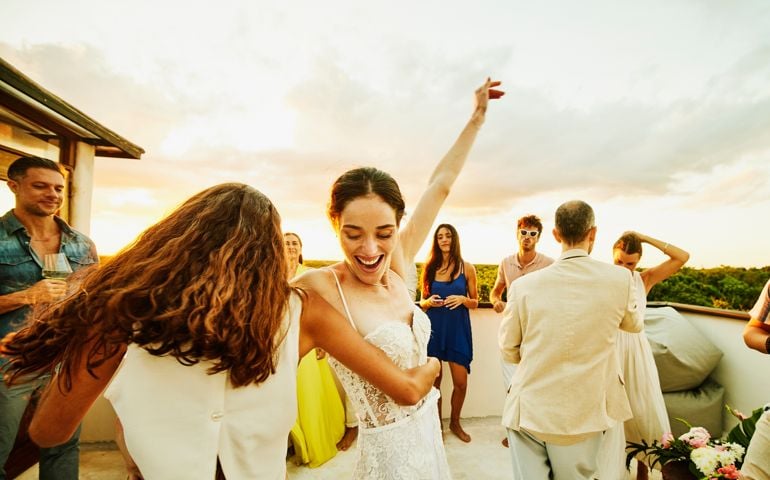 Medium wide shot of smiling bride and friend dancing during party on rooftop deck after