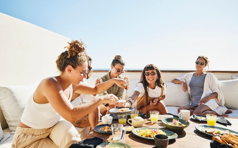 Medium wide shot of smiling female friends sharing breakfast on deck of luxury suite at tropical resort