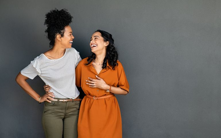 Two women friends embracing and laughing on grey backgroun