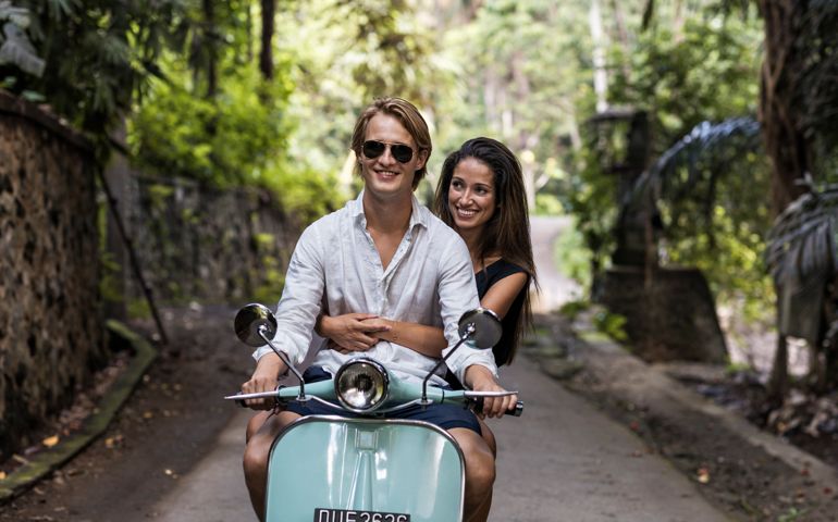 Young couple riding classic scooter along minor treelined road