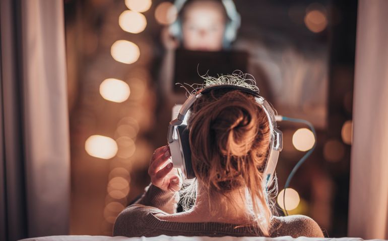Woman using tablet pc in evening wearing big headphones, sitting in front of a big window.
