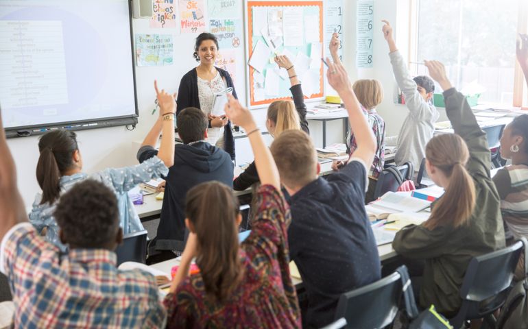 Rear view of teenage students raising hands in classroom