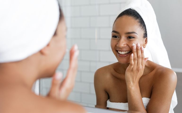 Happy woman putting on facial moisturizer with hand in bathroom mirror