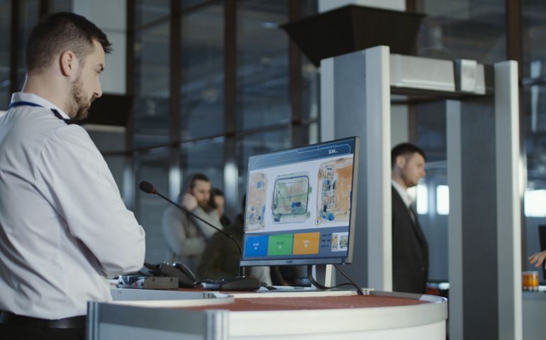 Man in uniform standing at counter at checking point and watching at monitor with x-ray of luggage.