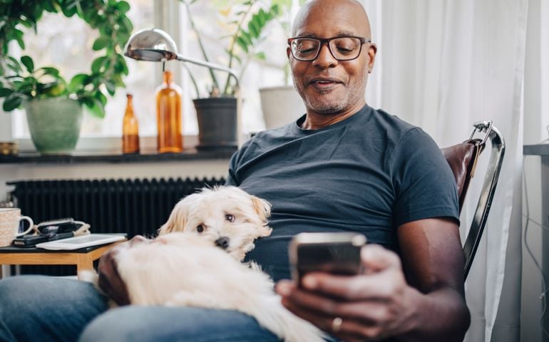 Smiling retired senior male using smart phone while sitting with dog in room at home