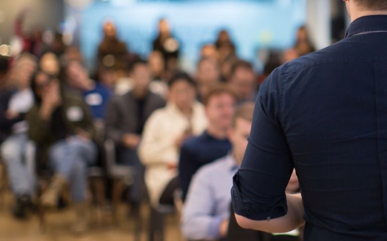 Conference photo audience and speaker giving speech. Seminar presenter on a panel during forum. Corporate manager in sales executive training discussion on stage. Investor pitch presentation.