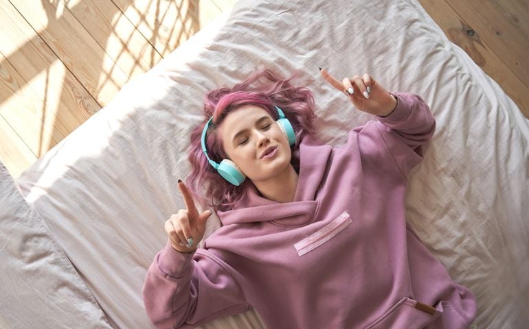Happy funny teen girl with pink hair wear headphones lying in comfortable bed listening new pop music enjoying singing song with eyes closed relaxing in cozy bedroom at home. Top view from above.