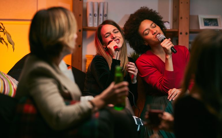 Happy multicultural friends sitting at home, laughing, singing, and having fun on karaoke night.
