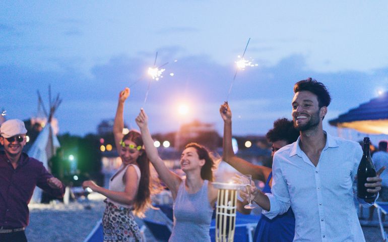 Happy friends making evening beach party outdoor with fireworks and drinking champagne - Young people having fun at chiringuito bar with dj set - Focus on right man face - Youth and summer concept