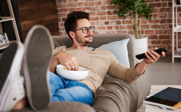 Content man lying on couch at home with bowl of potato chips watching TV