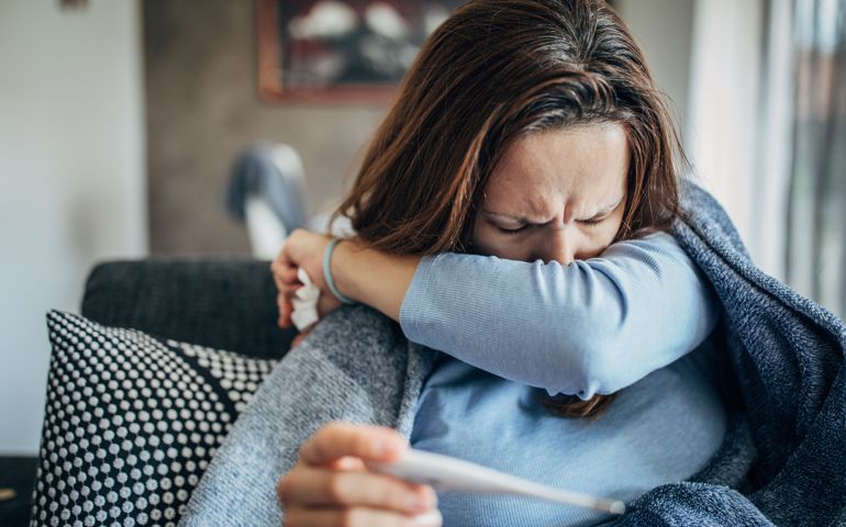 Woman with fever symptoms sitting on sofa and holding thermometer