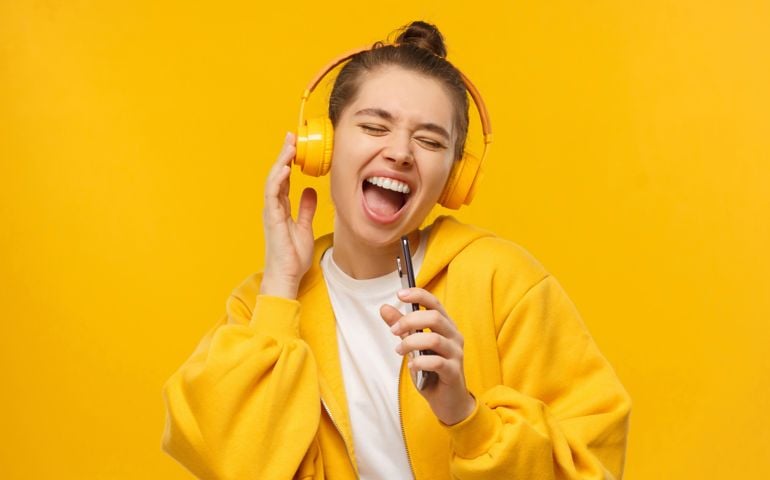 Trendy girl singing favorite song out loud in phone as mic, wearing wireless headphones, isolated on yellow background.