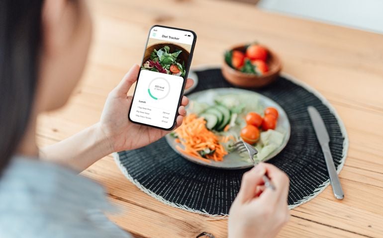 Young Woman Using Calorie Counter App On Smartphone While Eating Salad