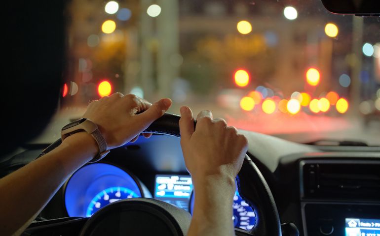 Close up of driver hands holding steering wheel driving car with blurred city street lights on background at night