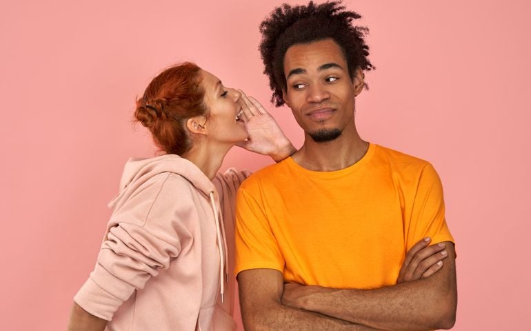 Ginger female whispers secret to Afro American boyfriend who has cheerful expression gossip together