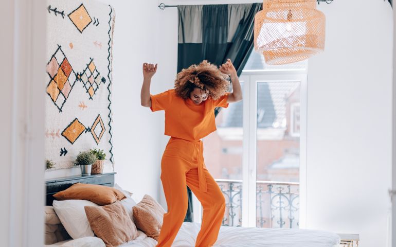happy young woman in orange outfit dancing on bed