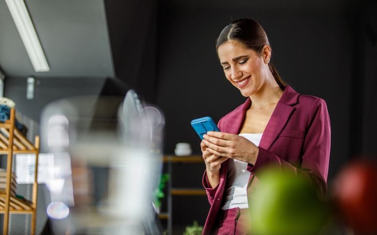 Young businesswoman sitting at the edge of her desk and responding to a text message via smart phone