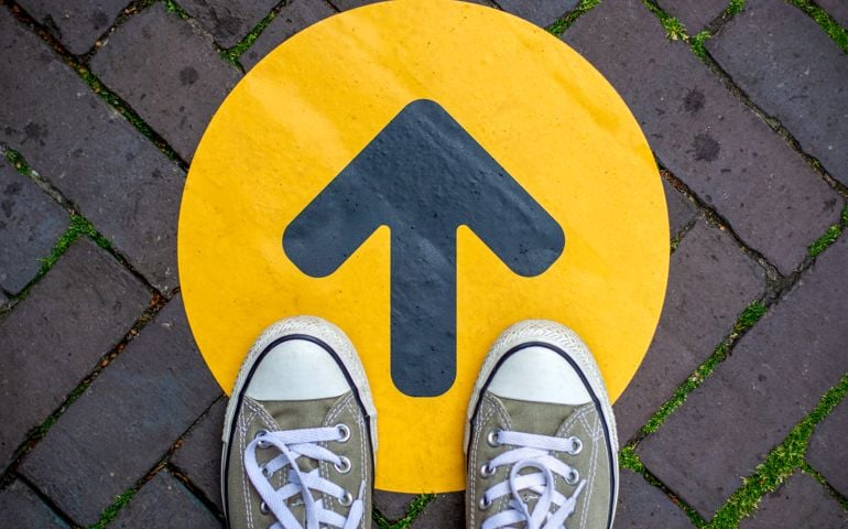 Feet wearing sneakers in front of arrow on road outdoors. Direction arrow in front of a store for the social distancing during the covid-19 pandemic.