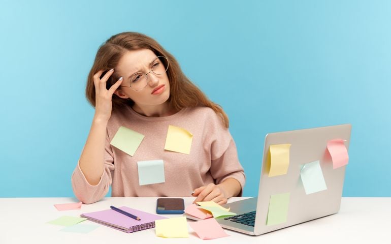 Pensive upset woman employee in nerd eyeglasses sitting at workplace office, all covered with sticky notes