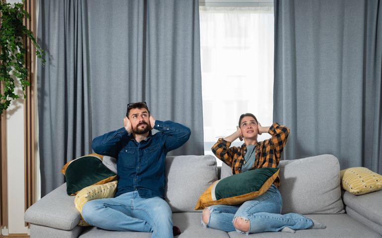 Young couple is sitting on a sofa in their apartment looking up and holding their hands to plug their ears as a neighbor upstairs is having a party and playing loud music or renovating the apartment