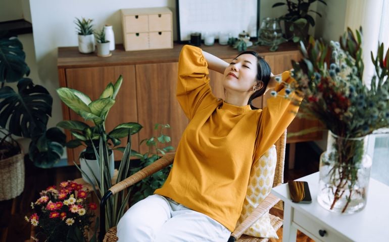 Young Asian woman with hands behind her head, taking a break and relaxing on armchair at cozy home, surrounded by green houseplants. Lifestyle, wellbeing and wellness concept