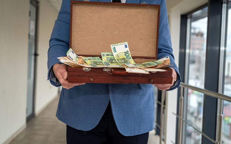 a man in suit stands with an open case in which a large amount of money