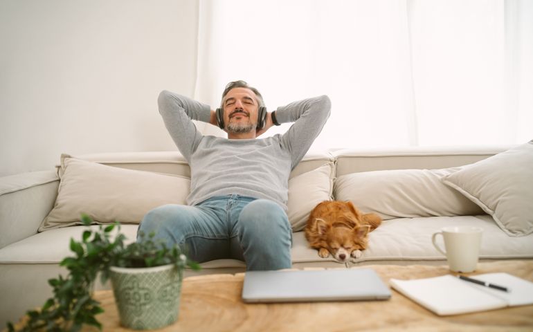 Calm Middle age Caucasian man sitting on sofa listening to music enjoying meditation for sleep and peaceful mind in wireless headphones, leaning back with his lovely chihuahua dog sit besides.