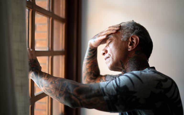 Worried mature man standing at home, close to a window, with hand in the head