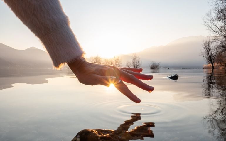 Detail of hand touching and caressing water surface of beautiful lake at sunset, mountain view. Purity freshness clean concept, one person touching lake with hand