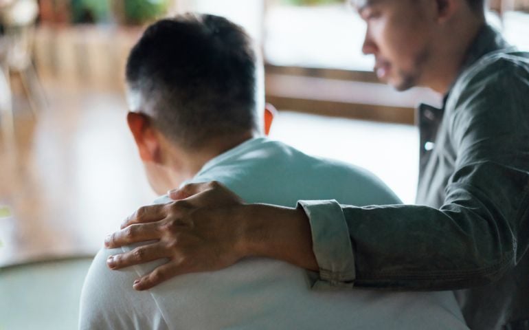Rear view of son and elderly father sitting together at home. Son caring for his father, putting hand on his shoulder, comforting and consoling him. Family love, bonding, care and confidence