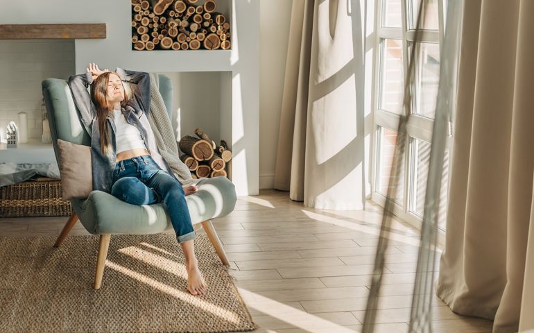 Young woman at home sitting on modern chair in front of window relaxing in her living room reading book and drinking coffee or tea