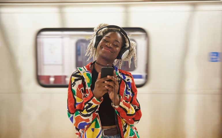Black woman listening to cell phone with headphones near subway
