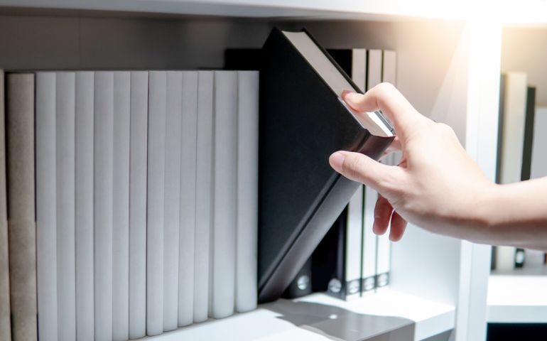 Male hand choosing and picking black book from white bookshelf in public library. Education research and self learning in university life concepts