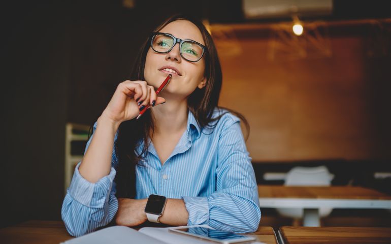 Dreamy woman podring while working on journalistic publication sitting with notebook in cafe,thoughtful female student in eyewear doing homework task solving problems and analyzing information
