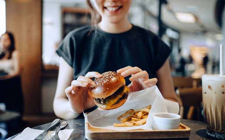 Smiling young Asian woman enjoying freshly made delicious burger with fries and a glass of iced coffee in a cafe