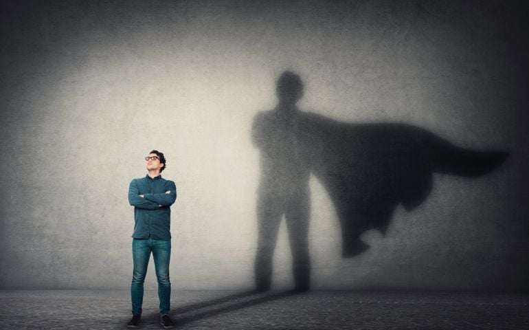 Brave man keeps arms crossed, looks confident, casting a superhero with cape shadow on the wall. Ambition and business success concept. Leadership hero power, motivation and inner strength symbol.