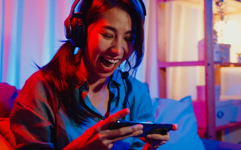 Happy asia girl gamer wear headphone competition video game online with smartphone excited talk with friend sit on couch in colorful neon lights living room at home.