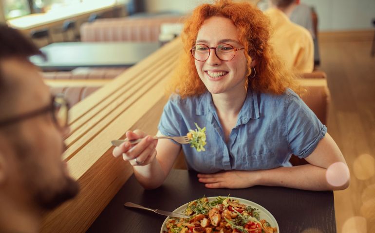 A close-up of a cheerful woman who enjoys hanging out with a friend and eating a Caesar salad.