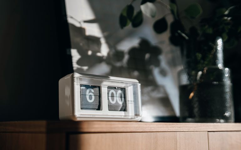 A white digital flip clock shows six o'clock on a brown wooden cabinet next to a fresh eucalyptus plant in the living room, with sunbeam shining through the window on a fresh beautiful morning. A brand new day, fresh start, fresh energy, new opportunities