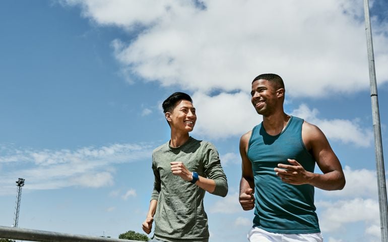 Active men jogging outdoors on blue cloudy sky with copy space. Two athletic guys or young sports friends running together, doing their routine cardio workout and fitness exercise in the city